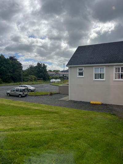 The image depicts a scene in Tyrrellspass, Ireland, on a cloudy day. In the foreground, a well-maintained green lawn slopes upwards. Beyond the lawn, a gravel parking area is visible, containing two parked cars: a silver minivan and a silver sedan. To the right, the side of a light-colored building with a dark, tiled roof is prominent, featuring white-framed windows. A bright yellow object, possibly a traffic barrier or a construction item, lies on the gravel near the building. In the background, a line of trees borders the parking area, and further back, several buildings are visible, including a large white cylindrical tank that appears to be a propane tank, and some houses with red roofs. A cellular tower stands tall in the distance. The sky is filled with a mix of white and gray clouds.