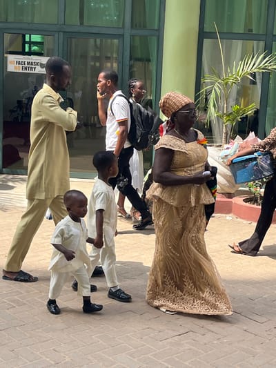 The image depicts a paved outdoor area in Madina, Ghana, beside a modern building with large green-framed glass doors and windows. The ground is covered with light brown interlocking bricks. Bright sunlight illuminates the scene, casting minimal shadows.
In the foreground, two young Black boys are walking towards the right, holding hands. Both are dressed in matching light cream-colored traditional two-piece outfits, consisting of short-sleeved tops with buttoned collars and matching trousers. The younger boy on the left wears black socks and shiny black dress shoes, while the older boy on the right wears black loafers.
To the left, a Black man in a light tan long-sleeved tunic and matching trousers, with dark sandals, stands with his arms crossed, looking towards the right. Behind the boys and to the left of the older woman, a young Black man walks right, wearing a white short-sleeved Ghana national football team jersey with a Black Stars logo, black trousers, and dark shoes. He carries a black backpack and holds a black smartphone to his right ear. A camera body hangs from his right hip.
In the mid-ground right, a mature Black woman walks right, dressed in an elaborate golden-tan traditional Ghanaian lace outfit (kaba and slit) with intricate embroidery and a peplum top. She wears glasses, a patterned brown headwrap, and a small Ghanaian flag pin on her chest. She holds a white object in her left hand and a bag with a red, yellow, and green strap in her right.
In the background, the building features a green vertical pillar on the right and a glass door with a white sign that reads "NO FACE MASK NO ENTRY" above "IMMEDIATE PARENTS' MINISTRY." A small potted palm tree is visible to the right of the green pillar. Partially visible on the far right is a person's lower body in dark trousers and sandals, carrying a blue woven plastic basket. Additional indistinct figures are visible through the reflective glass doors.