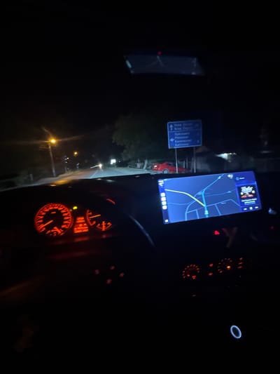 The image, taken from the interior of a car at night, shows the driver's perspective on a road. In the center of the view is a mobile phone displaying a map with a route highlighted in yellow. To the left, there is a road sign indicating directions to Nova Zorya, Preslaven, and Kalitinovo, indicating the location is in the municipality of Mogila, Bulgaria. The car's dashboard is partially visible, showing the speedometer, tachometer, and other instrument panel details. The ambient light is low, with streetlights visible in the distance outside the car.