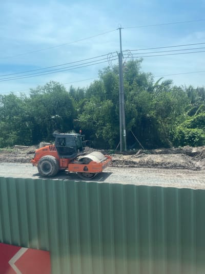 This image captures an outdoor scene with a construction site in Dong Nai City, Vietnam. In the foreground, a green corrugated metal fence runs horizontally across the bottom of the frame. Behind the fence, an orange road roller is positioned on a gravel surface. The road roller is a piece of heavy machinery with a large cylindrical drum at the front and tires at the back. It has the markings "QD-09" and "3414" visible on its body, along with a "HAMM" logo. The background features dense green foliage, including trees and palm fronds, suggesting a natural or semi-rural environment. A utility pole with wires stretches upwards into a partly cloudy blue sky. The ground around the road roller appears to be dirt and construction debris, indicating ongoing work.