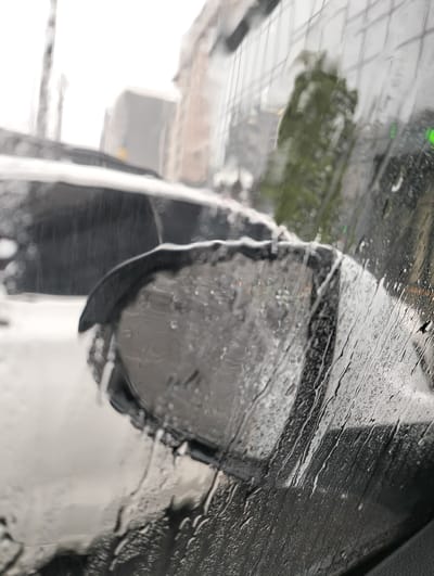 The image captures a view from inside a vehicle during inclement weather, likely rain or sleet. The foreground is dominated by the car's wet side window and rearview mirror, both heavily streaked and beaded with water droplets. The window surface shows numerous vertical rivulets of water, distorting the view beyond.

Through the rain-obscured glass, parts of the urban environment are visible. In the mid-ground, the car's exterior, possibly a white or light-colored body, is seen, with what appears to be a layer of snow or slush accumulated on its surface, particularly on the top of the side mirror housing and the adjacent part of the car body. This suggests a cold, wet environment, possibly a mix of rain and snow.

In the blurred background, several buildings are discernible, hinting at an urban setting in Ivano-Frankivsk, Ukraine. One building has a grid-like structure on its facade, possibly glass panels, reflecting some green foliage, likely a tree or shrub. Another building appears lighter-colored and more distant. The overall lighting suggests an overcast or gloomy daytime, characteristic of rainy or snowy weather. There are no visible people or discernible text in the image. The scene conveys a sense of a cold, wet, and active weather event from the perspective of someone inside a vehicle.
