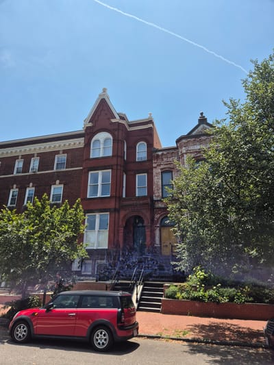 A bright, sunny day in Washington, D.C. features a row of brick townhouses. The foreground shows a red Mini Cooper Clubman parked on the street. A set of concrete stairs leads up to the entrance of the middle townhouse, which has a dark green door and ornate arched windows. Adjacent to this is another townhouse with a pale yellow door. Lush green trees and landscaping surround the buildings. A contrail streaks across the clear blue sky.