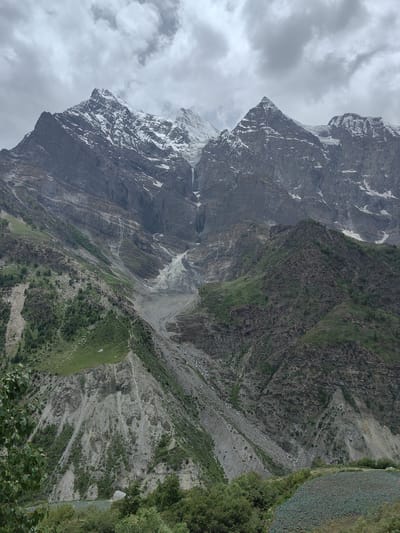 The image depicts a majestic mountain landscape under a cloudy sky. In the foreground, lush green vegetation and a terraced field of crops suggest human cultivation. The middle ground features steep, rocky slopes dotted with sparse trees and vegetation, with a prominent grey scree slope descending from above. The background is dominated by towering snow-capped mountains, their rugged peaks and vastness emphasizing the wild, natural beauty of the scene. A thin waterfall or stream is visible on the face of the distant mountains. The overall impression is one of a remote, natural environment where agriculture meets rugged, mountainous terrain. The location is identified as an unknown place in India.
