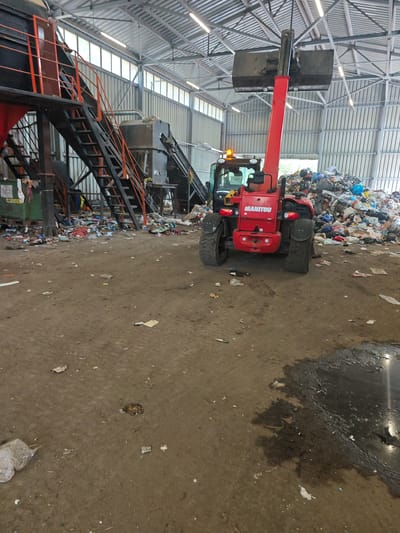 The image shows the interior of a waste processing facility, likely a recycling or sorting plant. A red "MANITOU" brand telehandler/loader is positioned in the center, its bucket raised, seemingly working with a large pile of mixed waste (plastic, paper, textiles, etc.). To the left is a large metal structure with stairs and conveyor belts, indicative of the sorting and processing equipment. The floor is dirty and littered with scattered waste. The building has a metal roof and walls, with natural light entering through windows along the upper walls. The image does not confirm the location in Barutin, Bulgaria.