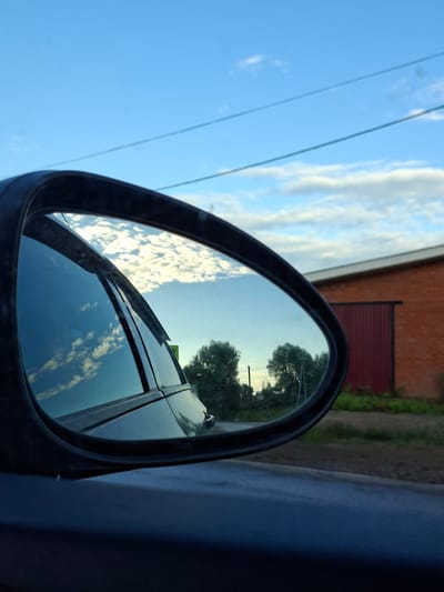 The image is a close-up shot of a car's side-view mirror, with the reflection showing a cloudy sky and trees. The mirror is framed by a black casing. In the foreground, outside the mirror's reflection, we see a blue surface, likely part of the car's body, and a portion of the car's windshield wiper.
The reflection in the mirror captures a bright blue sky dotted with fluffy white clouds. The clouds are illuminated from behind, suggesting it might be late afternoon or early evening, with the sun low in the sky. Silhouetted against the sky are the tops of several trees. The reflection also includes a portion of the car's roof and side window, which mirror the sky and clouds.
In the background, visible outside the mirror to the left, is a red brick building with a white trim and a red corrugated metal section. Green foliage is visible near the building, suggesting a rural or suburban setting. Two thin, dark lines, possibly power lines or wires, traverse the sky.
The overall impression is one of a serene moment, possibly a drive through the countryside. The lighting and weather suggest a pleasant day. No people are visible, and there is no discernable text in the image. The location context provided is Chumna, Russia, which aligns with the visual cues of a rural or semi-rural setting.