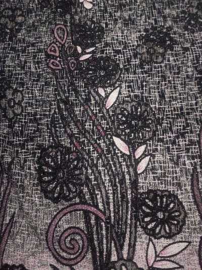 A close-up image shows a floral textile pattern. Black floral shapes are prominent, interspersed with vertical black lines. Small, pale leaves accent the flowers. The background has a textured, cross-hatched effect in gray tones. A purplish-pink spiral is visible towards the bottom. The overall pattern is dense and intricate.