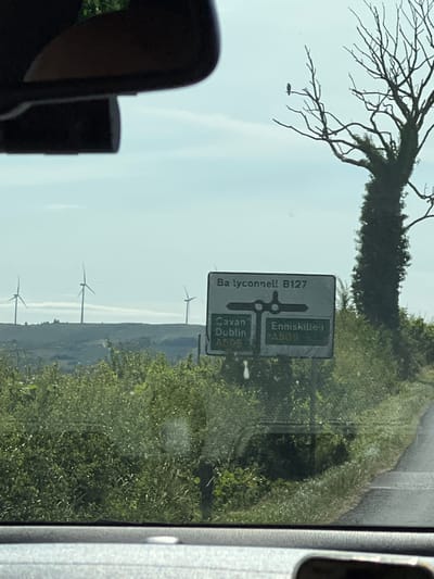 The image shows a road sign indicating directions to Ballyconnell B127, Cavan, Dublin, and Enniskillen A509. The sign is situated in a rural area with a tree to the right, wind turbines in the distance on a hilltop, and lush green vegetation. The sky is overcast. The perspective suggests the photo was taken from inside a vehicle, looking forward through the windshield. The scene is located on the outskirts of Derrylin, United Kingdom, likely on a road leading towards these destinations.