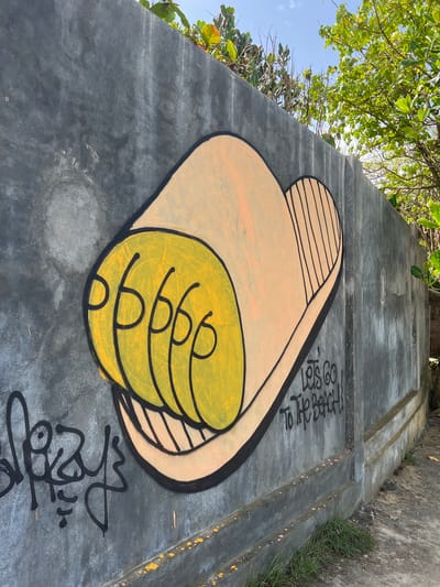 The image depicts a section of a grey concrete wall adorned with graffiti in Midigama, Sri Lanka. A prominent, large-scale graffiti artwork features a stylized yellow shape with black outlines, possibly representing a banana or a similar fruit, with curved lines suggestive of segments. To the right of this artwork, handwritten text reads "Lets GO TO The BEACH!". Below the central artwork and to the left, there is another piece of graffiti in black, stylized lettering. The wall itself shows signs of wear and texture. Green foliage from trees and shrubs is visible at the top of the wall, and the ground in front of the wall is paved concrete with some grass growing. The scene suggests an outdoor, possibly urban or coastal, environment.