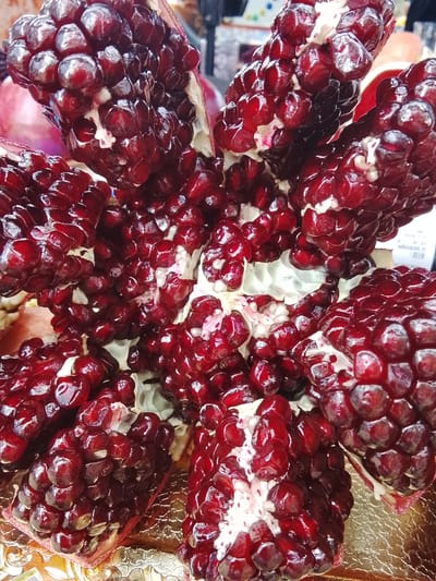 The media file is a close-up, overhead shot of a ripe pomegranate that has been opened and is displayed on a golden surface, possibly a tray or decorative plate. The pomegranate is segmented, revealing its numerous ruby-red arils, which are plump and glistening with juice. The arils are tightly packed within the fruit's white pith. Some arils show a deeper, almost blackish-red hue, indicating their ripeness. The overall impression is one of freshness and abundance. The background is slightly blurred, suggesting it might be a marketplace or a food display, with hints of other fruits and possibly some subtle text on a label in the far right. The lighting is bright, highlighting the glossy texture of the arils and creating small reflections. There are no people or animals visible in the media file. The setting appears to be indoors, given the controlled lighting. The time of day and weather conditions are not discernible from the image. The only visible text, though blurry, appears to be on a white label in the upper right portion of the image, with some Cyrillic characters faintly visible, consistent with a location in Russia.