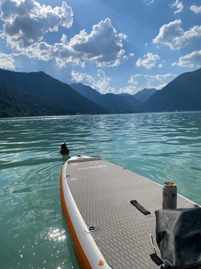 A low-angle, wide shot captures a paddleboard on a calm, turquoise lake. The paddleboard is positioned in the foreground, angled towards the right side of the frame, with its textured, grey surface and orange trim visible. A black bag and a silver can are resting on the paddleboard. In the distance, dark green mountains rise sharply against a bright blue sky filled with fluffy white clouds. The sun glints off the water's surface, creating sparkling highlights. A small, dark object, possibly a duck, is visible in the water near the paddleboard's edge. The image evokes a sense of peaceful recreation on a sunny day.