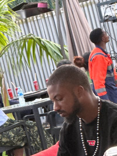 This media file depicts a scene with several people, likely in an outdoor setting. One individual in the foreground is wearing a black garment adorned with a double row of small, light-colored beads and a red emblem with a white "P". Another person, visible in the background, is wearing an orange jacket with blue and grey reflective stripes. The setting appears to be informal, with elements like corrugated metal structures and lush greenery, possibly indicating a rural or semi-urban environment in Rubuchi, Nigeria. There are also several red cylindrical objects with sticks, resembling popsicles, and a plastic water bottle visible. The orientation of the image is tilted. The time of day and weather are not discernible. No specific emotions are conveyed.