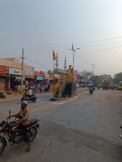 The image shows a street scene in Kothacheruvu, India. A statue stands on a platform in the middle of the road. Two orange flags are placed on the platform. Motorcycles are driving on the street, and people are walking on the sidewalks. Shops line the street. There is a mosque with two minarets in the background. The sky is overcast.