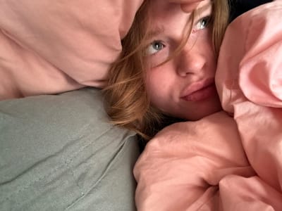 A close-up, high-angle image depicts a young individual with reddish-blonde hair and light-colored eyes, positioned amidst rumpled bedding in an indoor setting in Rosmalen, Netherlands. The person is lying down, with their gaze directed upwards and slightly towards the right side of the frame. Their left cheek rests against a volume of light pink fabric, likely a blanket or duvet. A segment of the person's right hand or fingers is visible near their forehead, lightly touching the hairline. The individual's light complexion is evident, featuring full lips. To the lower-left, a light grey-green textured fabric, possibly a shirt or another layer of bedding, is present. The overall lighting is soft and ambient.