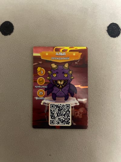 The image shows a close-up, top-down view of a collectible card lying on a textured, gray surface that appears to be a blanket or fabric. The card features a cartoonish purple monster with yellow eyes and horns, described as "Альп" (Alp) and "легендарний" (legendary). To the left of the monster are three circular icons with numbers and text: a lightning bolt with "3," an alien head, and "прибульці" (aliens). Below these is a circle with "25" and the text "номер" (number). A QR code is located below the monster. The background of the card depicts a fiery, molten landscape. The surface the card is on has two black circular smudges, one on the left and one on the right, which could be shadows or marks. There is no indication of the city of Berezhany, Ukraine, in the image.