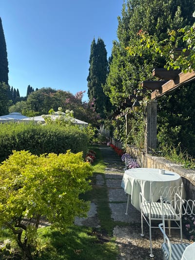 This is a sunny, daytime, exterior shot of a well-maintained garden with a stone pathway leading through it. In the foreground, a bright green, leafy bush is visible. To the right of the pathway, a round table covered with a light green tablecloth is set with a small white ashtray. Two white metal chairs with striped cushions are positioned at the table. A low stone wall runs alongside the pathway, topped with a wooden pergola structure covered in greenery. In the background, there are more trees, hedges, and two white tent-like structures. The location is identified as Maser, Italy.