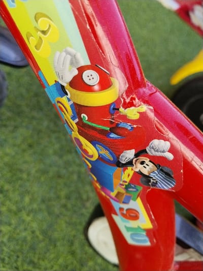 A close-up, angled shot depicts the mid-section of a red metal bicycle frame, likely a child's model, situated outdoors on green grass in Sucina, Spain. The frame features a large, rectangular, multi-colored decal. The decal primarily displays characters and elements from Disney's Mickey Mouse Clubhouse, including Mickey Mouse, a red cylindrical robot with a white button on its head, and a yellow wind-up key protruding from the robot's side. Other elements on the decal include geometric shapes and the numbers "9" and "10." The decal exhibits minor wear, with slight creases and lifting at the edges. The red paint of the bicycle frame shows several small scuffs and scratches. A portion of a white training wheel or stabilizer is visible at the bottom right, while out-of-focus green grass occupies the background, along with hints of another yellow object and a black tire.