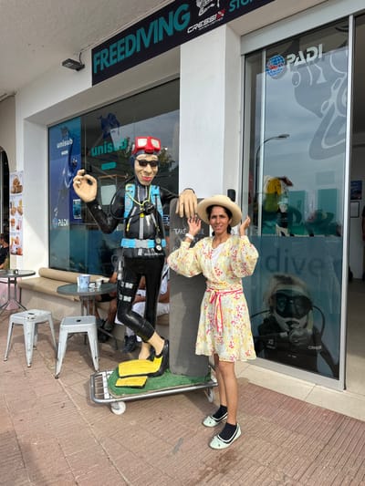 The image depicts an outdoor commercial street scene in L'estartit, Spain. An adult female is positioned centrally, to the right of a large diver mannequin. The female subject wears a straw hat, a yellow floral-patterned wrap dress with a pink sash, black ankle socks, and light blue ballet flats. Her hands are raised, palms outward, and she is smiling towards the camera. The caricature-style diver mannequin, dressed in a black wetsuit, red mask, black sunglasses, and yellow fins, stands on a wheeled cart to the female's left, with its right hand forming an 'OK' sign. The background consists of a storefront identified by a large 'FREEDIVING' sign and window decals for 'CRESSI DIVE CENTER,' 'PADI,' and 'unisub dive shop.' Reflections in the windows include a street light and a large image of a diver. To the far left, two light grey metal stools and a small round table with a dark cup suggest an adjacent cafe area. The ground surface is composed of reddish-brown paving stones.