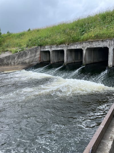 The image shows a concrete structure with multiple arched openings from which water is flowing. The structure is partially covered by a grassy embankment. The water in front of the openings is turbulent and foamy, indicating a significant flow. The sky is overcast. There is no visible human presence in the image. The scene is a section of a spillway or dam, likely part of a water management system.