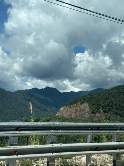 An eye-level, medium shot captures a scenic mountain landscape under a cloudy sky. In the foreground, a silver guardrail with reflective red and yellow markers runs horizontally across the frame. Behind the guardrail, tall, dry grass and some green vegetation are visible. The background features a dramatic mountain range covered in dense green foliage. A large, rocky cliff face is prominent on the right side of the mountains. The sky is filled with dramatic, grey clouds, with a small patch of blue visible in the upper center. Overhead, two dark power lines stretch across the top of the image. The scene appears to be captured from a moving vehicle on a road, likely in Nam Khanh Vinh, Vietnam, given the lush mountainous terrain and the presence of a guardrail common on roads in such regions. No people are present in the image.