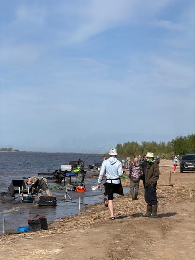 The image captures a fishing competition taking place on the shore of a body of water, likely in Krasnyi Bor, Russia. Multiple individuals are visible, equipped with fishing rods and gear, stationed along the shoreline. The setting includes a sandy bank transitioning into shallow water, where anglers are positioned. Fishing equipment such as rods, stands, and various containers are scattered around each person, indicating organized activity. The sky is clear and indicates daytime.