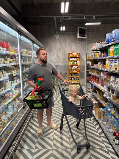 The image captures a detailed scene within a supermarket aisle in Sobinka, Russia. An adult male and a toddler are positioned in the foreground, engaged in grocery shopping.
The adult male, exhibiting a beard and short light-brown hair, is clad in a grey, distressed t-shirt, denim shorts, and light beige slip-on sandals. He stands with his left hand on the handle of a dark grey shopping cart, and his right hand holds a small black plastic shopping basket containing dark-wrapped items. A green cup with a red lid is also held in his right hand, positioned above the basket. His gaze is directed towards his right, past the refrigerated display.
A toddler, with curly blonde hair and wearing a light-colored t-shirt, is seated inside the main compartment of the dark grey shopping cart, facing towards the viewer's right. The toddler appears to be smiling, with both hands resting on the edge of the cart.
To the left of the scene, a long refrigerated display case with glass doors extends along the aisle. It is stocked with various dairy products, including yogurts and cheeses, and features numerous yellow price tags. To the right, multiple metal shelves are stocked with a variety of packaged groceries. Notable products include baby formula (Nestogen, Nutrition, Nestle), cereals, and milk products. Many shelves exhibit gaps, indicating sold items. The lowest shelf displays large clear bottles of water and smaller red-capped bottles. In the background, center-left, a tall, stacked display of "Lay's" potato chips and sunflower seed packets is visible. Mounted on the back wall, center-right, is a white industrial fan or heater unit labeled "VOLCANO."
The supermarket features an industrial aesthetic with dark grey, textured concrete walls and a dark ceiling crisscrossed with metal conduits and exposed lighting fixtures. The floor is tiled with a distinctive chevron pattern in alternating shades of dark and light grey.