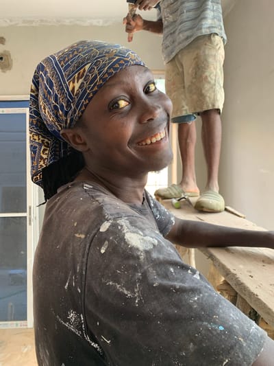 The photograph captures a close-up view of a person wearing a blue and yellow patterned headwrap and a dark t-shirt splattered with white paint. The person is smiling directly at the camera, their eyes open and looking towards the viewer. Their skin is dark, and their lips are slightly parted, revealing white teeth. In the background, another person, whose face is not visible, stands on a wooden platform. This second individual is wearing a horizontally striped t-shirt and light-colored shorts, also speckled with paint, and appears to be engaged in some activity involving a tool held in their right hand, possibly painting. The environment suggests an indoor construction or renovation site. The wall behind them is light-colored, and parts of a window frame are visible on the left. The lighting appears to be natural, coming from the right. This scene is located in Zawan, Nigeria, a city known for its construction and development projects.