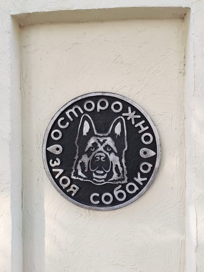 The image shows a close-up, eye-level shot of a circular metal plaque mounted on a textured, cream-colored wall. The plaque is black with raised silver lettering and an embossed image of a dog's head in the center. The lettering appears to be in Cyrillic script, arranged around the dog's head. The dog has a serious expression, with prominent ears and facial markings. The wall has a rough, stucco-like finish. The plaque is casting a subtle shadow on the wall. The scene appears to be outdoors, likely in a public space, given the mounting on a wall. The location is identified as Vasil'ki, Russia.
