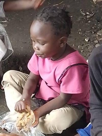 An African child, likely a girl, with dark skin and tightly curled black hair, is seated on the ground in Otulu, Nigeria. She is wearing a bright pink short-sleeved shirt with black trim along the shoulders and neckline, and light-colored trousers that appear worn and torn at the knees. The child is actively engaged in eating, using her hands to scoop food from a pile on a plastic sheet laid out on the dirt ground. The food is a light brown, lumpy substance. The environment appears to be outdoors, with the ground composed of dark earth mixed with small stones and debris. The child is positioned next to at least two other individuals, whose clothing is visible but their faces are not. One person on the left is wearing a light-colored garment, and another on the right is wearing a dark gray garment. The overall scene depicts a moment of communal eating in what seems to be a simple, outdoor setting.