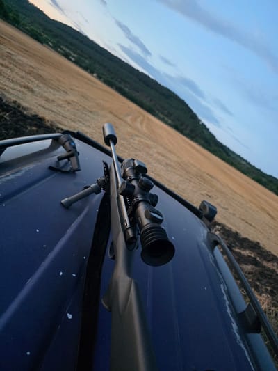 This media file shows a rifle with a scope and suppressor resting on the hood of a dark blue vehicle. A bipod is attached to the rifle. In the background, a harvested field stretches towards a tree-covered hillside under a pale blue sky with some clouds, suggesting either dawn or dusk. The sun is low, casting a soft light. The overall mood is serene and still, with a sense of preparedness. The location is identified as Levski, Bulgaria. No people are visible in the image.