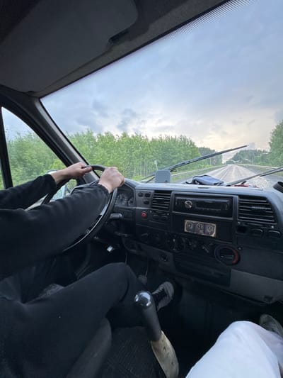 The image shows the interior of a van, viewed from the passenger's seat, looking forward. A person's hands are on the steering wheel, indicating they are driving. The dashboard is visible, with a radio, climate controls, and other indicators. The windshield shows a wet road and trees lining both sides, suggesting it is raining or has recently rained. The sky is overcast. The scene appears to be on a road outside of Pesterevo, Russia.