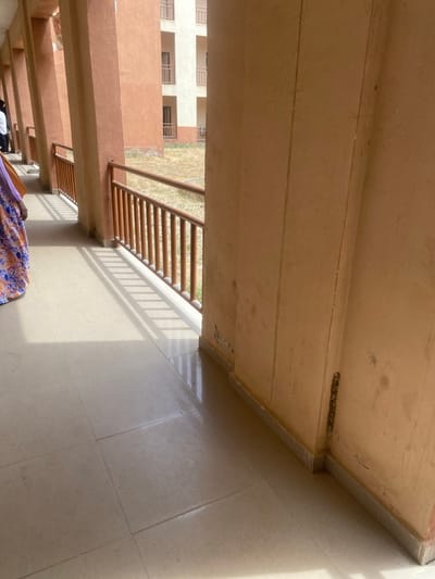 The image depicts a semi-enclosed outdoor corridor within a building complex in Baganakwo, Nigeria. The foreground features a light-colored tiled floor illuminated by strong sunlight, casting distinct parallel shadows from a balustrade. To the immediate right, a thick, peach-colored concrete pillar shows signs of wear and minor surface damage near its base, including an exposed dark vertical element and some paint loss. A light gray skirting tile runs along its bottom edge.
Along the left side of the corridor, a series of similar peach-colored concrete pillars forms an arcade, with a wooden or wood-toned metal balustrade and handrail positioned between them. Beyond this railing lies an open, undeveloped plot of land covered in dry grass and weeds. In the background, two multi-story buildings with peach and light beige facades are visible, featuring multiple windows and some balconies with railings.
One individual is partially visible on the far left, wearing a long patterned skirt or wrapper with blue, orange, and white floral or abstract designs. Another individual, primarily obscured by a pillar further down the corridor, is seen wearing a white shirt. No specific actions or interactions are discernible beyond presence in the corridor.