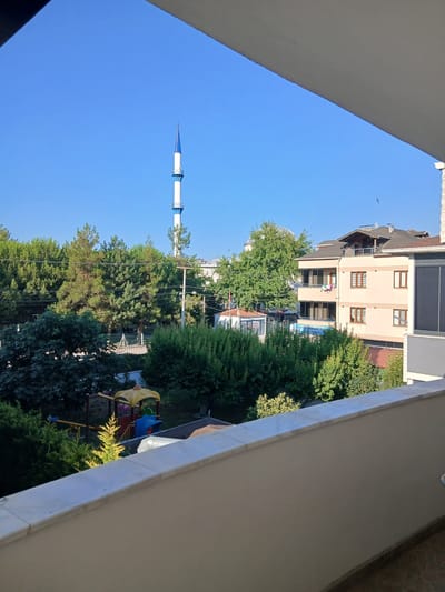 An exterior, eye-level, medium shot captures a sunlit, residential cityscape in İnegöl, Turkey. The foreground is dominated by a beige balcony wall with a marble coping. Beyond the balcony, a lush green landscape unfolds, featuring various trees, some with dense foliage and others with more sparse, needle-like leaves. A small, colorful playground with yellow, red, and blue elements is visible amidst the greenery. In the mid-ground, several buildings are present. One is a light beige, multi-story apartment building with brown-framed windows. Another building, further to the left, has a white-domed roof and is partially obscured by a large green tree. Towering above the scene in the background is a slender minaret of a mosque, white with blue accents, reaching towards a clear, vibrant blue sky. A few birds are discernible in the sky. Power lines traverse the middle ground, strung between utility poles. The scene appears to be a residential area on a clear, sunny day.