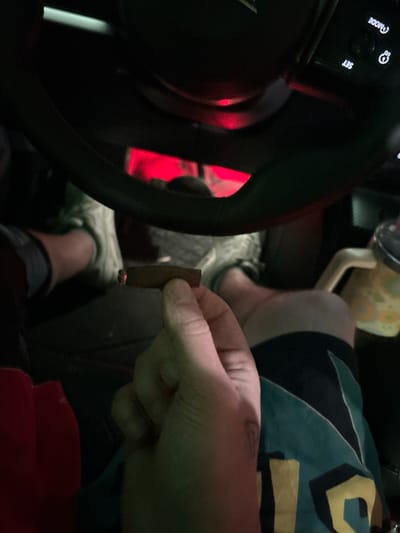 The scene depicts the interior of a vehicle in Reno, United States of America, characterized by low light conditions and a prominent red ambient light source originating from the dashboard or console area. In the foreground, a left hand holds a lit, partially consumed cigar or blunt, with its tip glowing orange. A faint tattoo is visible on the side of the hand/wrist. The subject's lap is visible below, covered by shorts featuring a teal, black, and yellow geometric pattern. A light-colored athletic shoe is present on the subject's foot. The lower portion of a black steering wheel is in the upper frame, displaying illuminated white controls labeled "SET" and "MODE". To the right, a light-colored beverage cup with a handle and dark lid is visible.