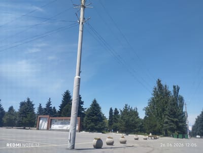 This media file depicts an outdoor scene on a clear day with a bright blue sky. In the foreground, a tall, light-colored utility pole stands prominently, with multiple wires extending from it across the sky. The ground is paved, resembling a parking lot or public square, marked with yellow lines.
To the left, a large, modern monument or information display is visible, featuring a map-like graphic and text in Cyrillic script. Surrounding this monument and stretching into the background are numerous evergreen trees, creating a lush, natural backdrop against the urban setting.
Several round, concrete planters with some foliage are placed in a row across the paved area, adding decorative elements to the scene. In the distance, a few people can be seen walking, suggesting a public space that is in use. The overall atmosphere appears calm and serene.
Visible text includes "REDMI NOTE 12" in the lower left corner and a date and time stamp "26.06.2026 12:05" in the lower right corner. The time stamp indicates it is midday. The setting is likely a public park or square in Karakol City, Kyrgyzstan, given the trees and monument.