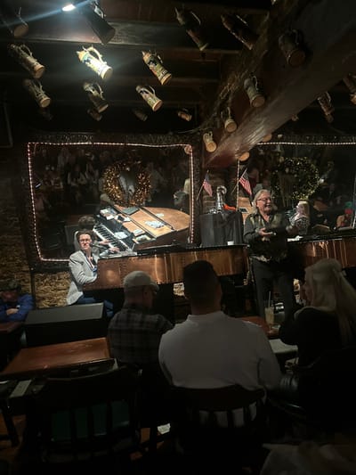 This image depicts a lively indoor scene within a bar, specifically **Pat O'Brien's Piano Bar in the French Quarter of New Orleans, United States**.
The environment is dimly lit, characterized by exposed dark brick walls and heavy wooden beams on the ceiling, from which numerous ornate ceramic beer steins are suspended as decor. A performance area is visible, featuring a large, unique instrument with copper paneling and riveted accents, equipped with two keyboards and multiple levers, likely an electronic organ or piano. Several US dollar bills are scattered across the top of this instrument.
Two male performers are central to the scene. On the left, a man wearing a light grey jacket and glasses is seated at the instrument, operating the keyboards. To his right, a second man with long, light-colored hair and glasses, dressed in a patterned vest over a dark shirt, stands and appears to be speaking or interacting with the audience, holding a dark, indistinct object in his left hand. A microphone stand is positioned between the two performers. To the far right of the stage, a small American flag on a pole is visible, alongside a silver, trophy-like object resting on a dark draped stand.
Behind the performers, a large rectangular mirror, framed by warm white string lights, reflects a seated audience, indicating a crowded venue. In the foreground, several patrons are seated at dark wooden tables with chairs, facing the stage. One man with a light-colored cap and plaid shirt is seen from behind on the left. In the immediate foreground, another man wearing a white long-sleeved shirt is seen from the back, seated next to a woman with long blonde hair holding a clear glass with a beverage and straw. The overall atmosphere suggests an engaging live music performance in a historic, bustling bar setting.