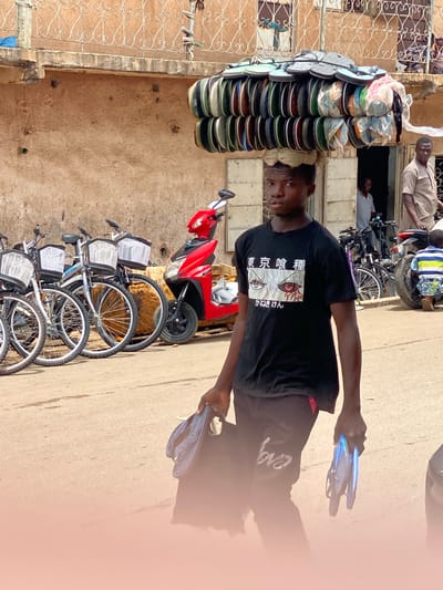 In Tudun Nupawa, Nigeria, a young man stands on a street alongside bicycles and a red scooter. He holds two pairs of flip-flops. A stack of flip-flops sits outside a building. People are visible through windows in the background.