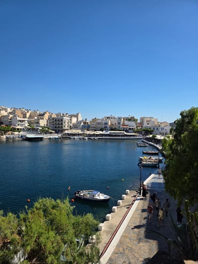 This is an image of a canal in Agios Nikolaos, Greece, with buildings lining one side and a paved walkway on the other. The water is a deep blue, and several boats are moored along the canal. A few people are walking on the walkway, and some are sitting at outdoor tables. The buildings are a mix of white and cream, with red-tiled roofs. There are trees and other greenery along the walkway. The sky is a clear blue, and the sun appears to be shining brightly, suggesting it's a warm, sunny day. The scene has a relaxed, leisurely atmosphere, indicating a typical day in a Mediterranean town. There is no visible text in the image.