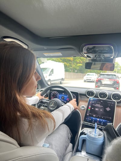 The image captures the interior of a Mercedes-Benz vehicle from the front passenger seat, facing forward. A female driver is seen in profile, with long, light brown hair, wearing a white long-sleeved top and grey pants. Her hands are positioned on the steering wheel, which bears the Mercedes-Benz logo. The dashboard features a digital instrument cluster and a large, vertically oriented infotainment screen displaying music playback (labeled "It's Up" by Savage) and Apple CarPlay interface elements. A light blue insulated cup with a straw is placed in a cup holder in the center console.
Through the windshield, a multi-lane paved road is visible under daylight conditions. Traffic includes a white commercial van in the adjacent lane to the left, and a white sedan followed by a dark SUV further ahead in the same direction of travel. Green trees and foliage are present along the right side of the road, behind a grey barrier or guardrail. The driver's eyes and forehead are partially visible in the reflection of the rearview mirror. The scene depicts a typical driving environment consistent with a road in the city of Newton, United States.