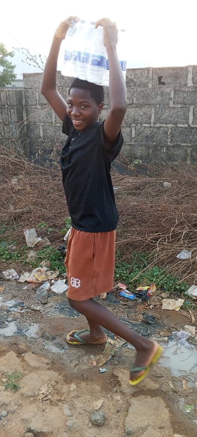 This image, captured in the city of Nassarawa, Nigeria, depicts an adolescent female in an outdoor, unpaved environment. The subject, with dark skin and short dark hair, wears a black short-sleeved polo shirt, brown shorts featuring a white "BB" logo on the left leg, and yellow and green flip-flops. She is shown in mid-stride, carrying a plastic-wrapped package containing multiple clear bottled water units balanced on her head, stabilized by both hands raised overhead. Her face displays a smile, directed towards the viewer. The ground consists of uneven dirt, marked by muddy puddles and scattered litter, including torn paper, plastic wrappers, and a small toy car. Behind the subject, a dense accumulation of dry, tangled brown vegetation is present, backed by a gray, textured concrete block wall. The upper background shows a bright, hazy sky with faint tree branches visible on the left.