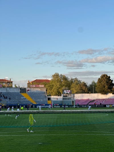 Here is a description of the image:
The image shows a soccer game taking place at Estádio Municipal Eng. Manuel Branco Teixeira in Chaves, Portugal. The stadium is partially visible, including the seating area, scoreboard, and advertising boards. The game is in progress, with a goalkeeper attempting to make a save in front of the net. Other players are visible on the field. The sky is blue with some clouds, suggesting it is a daytime game. There are logos from Haier and Hyundai.