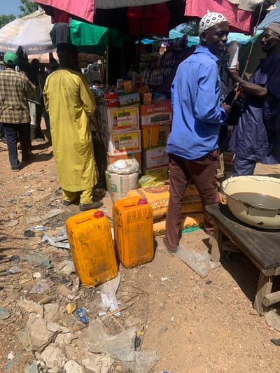 In Boi A, Nigeria, a bustling outdoor market scene unfolds under a canopy of colorful tarps. The ground is a mix of dirt and scattered debris, with plastic bags, wrappers, and other detritus.

In the foreground, two large, bright yellow plastic containers with red caps are prominently displayed. To their right, a large, rustic wooden table holds a metal basin filled with a dark liquid.

Several individuals are present, engaged in various activities. A man in a blue long-sleeved shirt and brown pants, wearing a distinctive white and black patterned cap, is holding a white bottle and appears to be in conversation with another man dressed in a dark blue robe, who is also holding a white bottle. Behind them, another man in a yellow robe stands with his back to the camera. Further back, other figures are visible, including a person in a plaid shirt and green cap.

The market stalls are laden with goods, including stacks of boxed items, suggesting a variety of products for sale. The overall atmosphere is one of commerce and daily life in a Nigerian market.