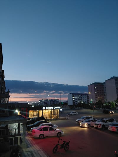 The image depicts a twilight scene in a city, likely Aktau, Kazakhstan, with several apartment buildings visible in the background. In the foreground, a commercial establishment with a prominent "GLOBAL COFFEE" sign is illuminated. A row of parked cars lines the street in front of the coffee shop and continuing down the road. Among the vehicles, a white sedan is parked closest to the viewer, and further down, a white SUV and other cars are visible. A bicycle is parked on the sidewalk. The sky displays a gradient of deep blue to a soft pink and orange hue near the horizon, indicating the sunset or twilight hour. Several streetlights are on, casting a warm glow on the surroundings. There are no prominent human figures captured in the frame, though a few people are faintly visible in the distance near the coffee shop and on the street. The overall environment suggests an urban setting at dusk, with commercial and residential structures coexisting.