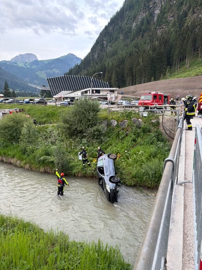 The image depicts a car accident scene in Campitello di Fassa, Italy, near a building with a distinctive modern architectural design, likely a sports or cultural facility. A black car has overturned and is partially submerged in a shallow, rocky river. Several firefighters in full protective gear are present, engaged in a rescue operation. One firefighter stands in the river, facing away from the camera, with their arm raised. Another firefighter is positioned on the riverbank, near the car, and two more are on a bridge above the scene, observing. A red fire truck is parked on the road behind them, and several other vehicles are visible in the background. The environment is mountainous, with a steep, tree-covered slope rising behind the road and buildings. The sky is overcast.