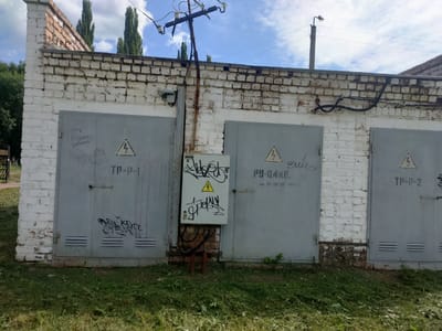 The image shows a section of a white brick building with three gray metal garage doors. The building is located in the city of Salavat, Russia. The doors are weathered and have various graffiti tags on them. The first door on the left has a heart symbol and some illegible writing. The middle door has a yellow triangular caution sign with a lightning bolt, and the text "РУ-0.4КВ." and "тел. 35-20-28." above it, along with more graffiti. The third door on the right has a similar caution sign and the text "TP-P-2". A small electrical box is mounted on the wall between the first two doors, also covered in graffiti. Electrical wires and poles are visible above the doors and the building. The ground in front of the doors is covered in grass and some debris. The sky is partly cloudy and blue.