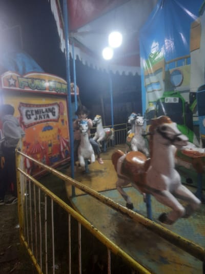 This image, captured in Sukasari, Indonesia, presents a nighttime view of an operational carousel at an outdoor fun fair. A worn yellow metal railing with vertical white bars defines the immediate foreground. To the left, a person, viewed from behind in a light gray hooded sweatshirt and dark pants, stands near a vibrant sign displaying "FUN FAIR" at the top and "GEMILANG JAYA" below, framed by carnival-themed illustrations on an orange background.

The carousel's platform is yellow with areas of blue discoloration from wear, supported by blue vertical poles that extend to a red and white scalloped canopy. Two prominent, bright circular lights illuminate the central area from above. A child, wearing a pink top, is seated on a white carousel horse in the mid-ground, facing forward. Another white and brown carousel horse, showing motion blur indicative of movement, is prominently positioned in the foreground. In the background, a large blue tarp or banner features a painted design resembling a building or ship, with green and brown elements including a visible porthole. The ground surrounding the carousel, visible beyond the railing, appears as dark, unpaved earth with scattered debris.