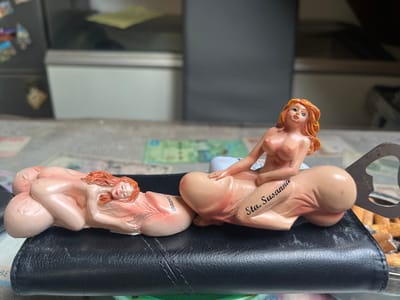 The image displays two small, suggestive figurines. The figurines are made of a beige-colored material with pinkish tones and depict nude women with red hair in stylized poses. The woman on the right is sitting atop a phallic-shaped object, with her legs spread. Text is visible on this phallic object, reading "Sta. Susanna." The woman on the left is lying down, seemingly resting on a similar phallic object. Both figurines appear to be part of a novelty item, possibly a bottle opener, as a metal opener is visible attached to the object on the right. The figurines are placed on a black, textured surface, possibly leather, with some paper currency or cards visible beneath them. The background is blurred, showing what appears to be a counter or shelf with drawers and some out-of-focus items. The lighting suggests it is daytime. There are no clear indications of weather or specific emotions conveyed by the figurines. The context provided places this scene in Schwäbisch Hall, Germany.