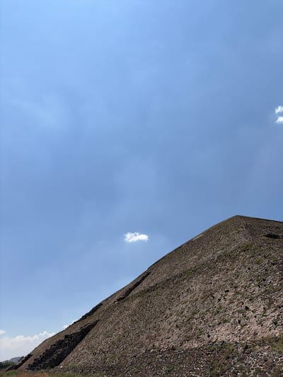 The image depicts a large stone structure or pyramid, likely part of the Teotihuacan archaeological site, as San Sebastián Xolalpa is a locality within the Teotihuacan municipality. The structure is composed of dark-colored stones, possibly volcanic rock, and exhibits signs of weathering or age, with some vegetation visible within the stone crevices. It dominates the right side of the frame.
To the left, a blue sky with sparse, small, white clouds fills the majority of the image. The sky appears clear and the lighting suggests it is daytime. The photograph is taken from a low angle, looking upward towards the structure and the sky. The angle makes the structure appear imposing.
