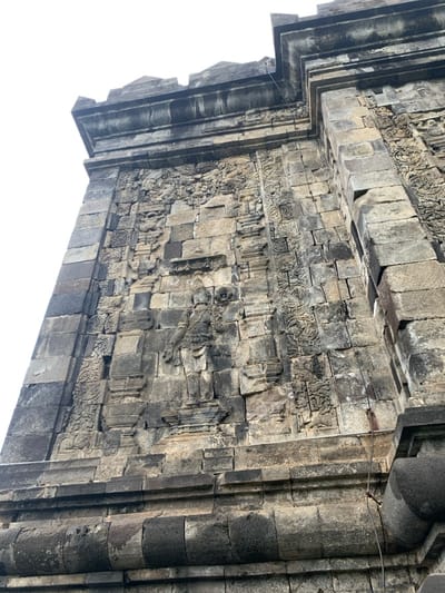 The image shows a close-up of a stone wall, the exterior of a temple in Mungkid, Indonesia, located at latitude -7.605 and longitude 110.23. The wall is made of large, rectangular stones, and is adorned with intricate carvings depicting figures and flora. The carvings are weathered and worn, indicating the age of the temple. The perspective of the image is from a low angle, looking up at the wall, which gives the impression of grandeur and age.