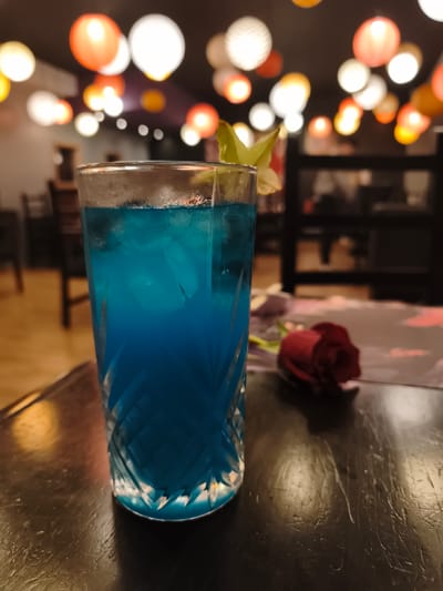 The image captures a tall, patterned glass filled with a bright blue beverage, likely a cocktail, containing ice and garnished with a star fruit slice. The glass is placed on a dark wooden surface that shows signs of wear. A single red rose lies next to the glass. The background suggests an interior setting, possibly a bar or restaurant, with a variety of illuminated, round lanterns hanging from the ceiling, creating a warm, atmospheric glow. Chairs and tables can be seen in the background, indicating a dining area. Based on the presence of star fruit, a tropical fruit, and the style of the dining establishment, it is plausible that this scene could be in Quito, Ecuador, given its equatorial location and diverse culinary offerings.