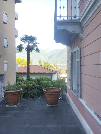 The image depicts an outdoor scene with a balcony in the foreground. Two large, terra cotta planters sit on a dark grey, textured surface, possibly a terrace or rooftop. The planter on the left contains sparse, green vegetation. Behind the planters, a black metal railing encloses the space, and beyond that, a dense line of green foliage.
In the mid-ground, a tall palm tree stands prominently. Behind the palm tree, a building with a terracotta-tiled roof is visible, along with other houses and trees. The background reveals rolling hills and mountains, suggesting a mountainous or hilly landscape. The sky is bright and appears to be a clear, pale blue, indicating daytime.
To the right of the image, a portion of a building with pinkish-orange siding is visible. This building has a balcony with an ornate railing and a window with closed blue shutters. Architectural details like decorative trim and a stone bracket supporting the balcony are also apparent. The perspective suggests the photo was taken from a balcony or elevated vantage point, looking outwards and slightly downwards.
The overall impression is a tranquil, scenic view from a residential area, possibly a vacation spot, given the palm tree and mountainous backdrop. The lighting suggests it might be morning or late afternoon, with the sun high in the sky. There is no visible text in the image.